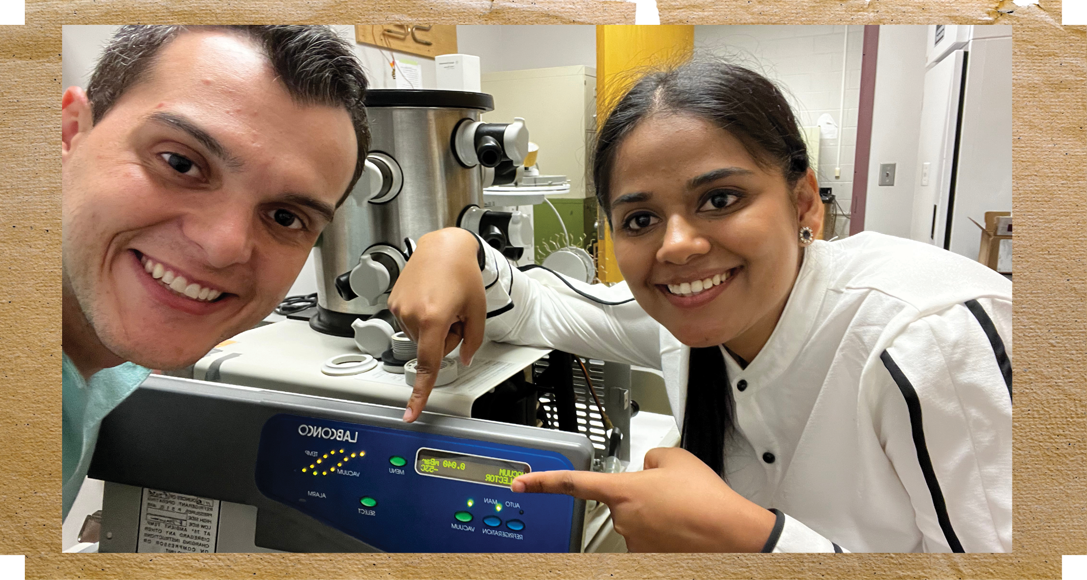 Dr. Eduardo Calixto is on the left side with a huge smile and on the left is Jasleen Kaur, also with a huge smile, pointing at the screen of the feeze dryer that separtes both herself and Eduardo.